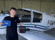 Un jeune pilote de l'Aéroclub de l'Aisne tout juste diplômé, tenant son carnet de vol PPL devant un avion de tourisme dans un hangar.