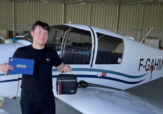Un jeune pilote de l'Aéroclub de l'Aisne tout juste diplômé, tenant son carnet de vol PPL devant un avion de tourisme dans un hangar.