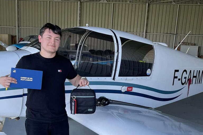 Un jeune pilote de l'Aéroclub de l'Aisne tout juste diplômé, tenant son carnet de vol PPL devant un avion de tourisme dans un hangar.