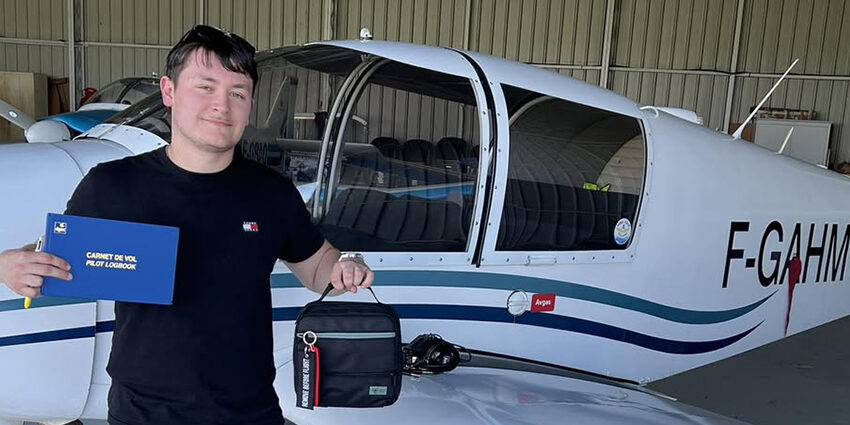 Un jeune pilote de l'Aéroclub de l'Aisne tout juste diplômé, tenant son carnet de vol PPL devant un avion de tourisme dans un hangar.