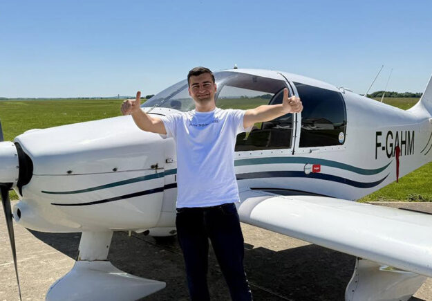 Le jeune pilote Serdem souriant, faisant un double pouce levé devant un avion de tourisme blanc immatriculé F-GAHM sur le tarmac ensoleillé de l'Aéroclub de l'Aisne.