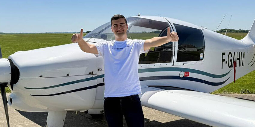 Le jeune pilote Serdem souriant, faisant un double pouce levé devant un avion de tourisme blanc immatriculé F-GAHM sur le tarmac ensoleillé de l'Aéroclub de l'Aisne.